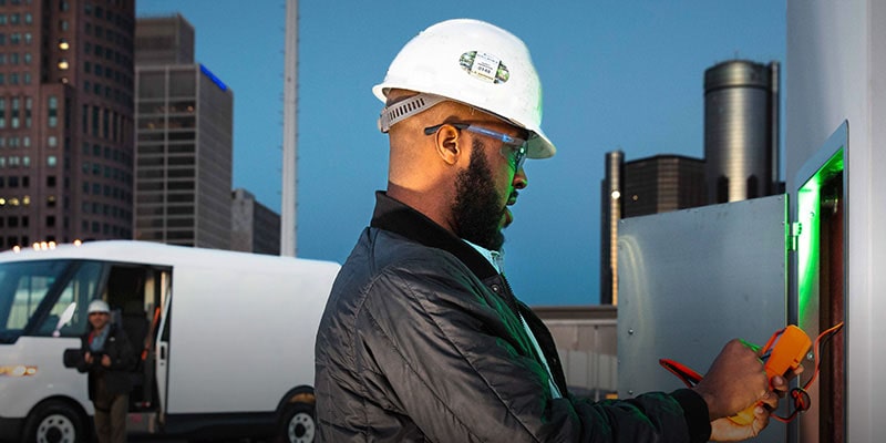 A service technician checking an electrical panel with their Brightdrop in the background