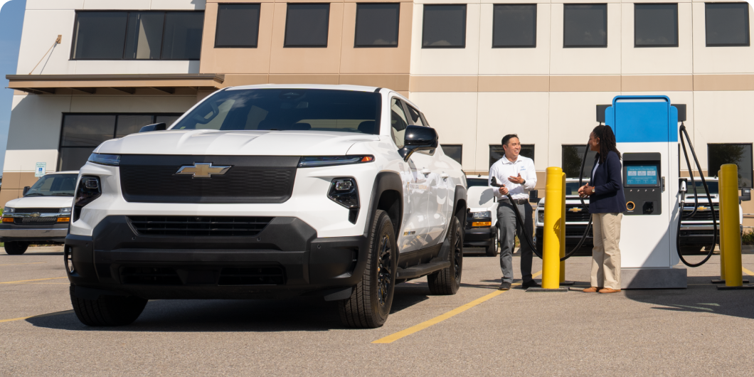 A Silverado EV plugged in at a charging station, while two people talk to each other. 