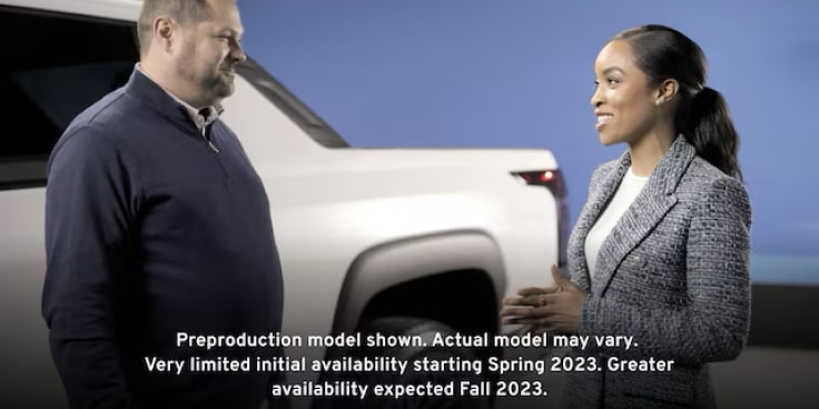 A man and woman stand next to the exterior of the Summit White 2024 Silverado EV discussing its capabilities.