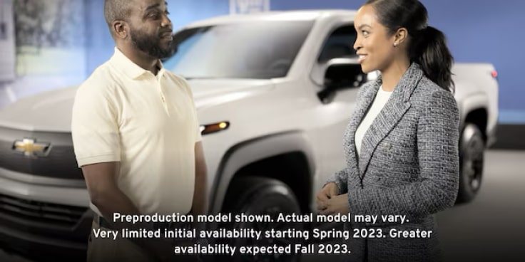A man and woman stand next to the exterior of the Summit White 2024 Silverado EV discussing its capabilities.