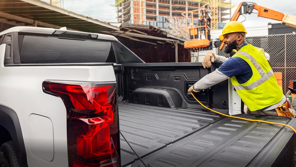 An Exterior view of a Construction Worker connecting a charger cable within the trunk area of the Silverado EV at a Construction Site.