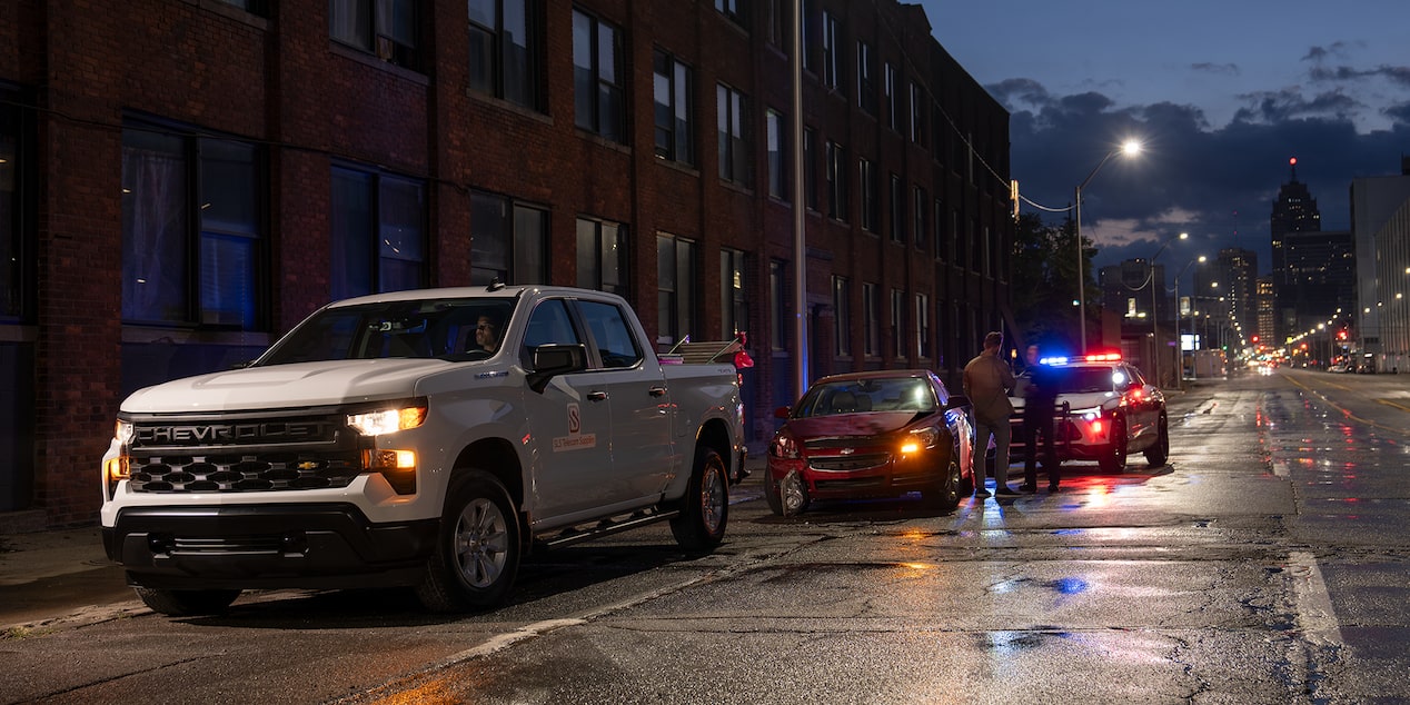 A white Chevrolet Silverado parked on a rainy street at night. A cop in the distance assists a vehicle that's been in a car crash.