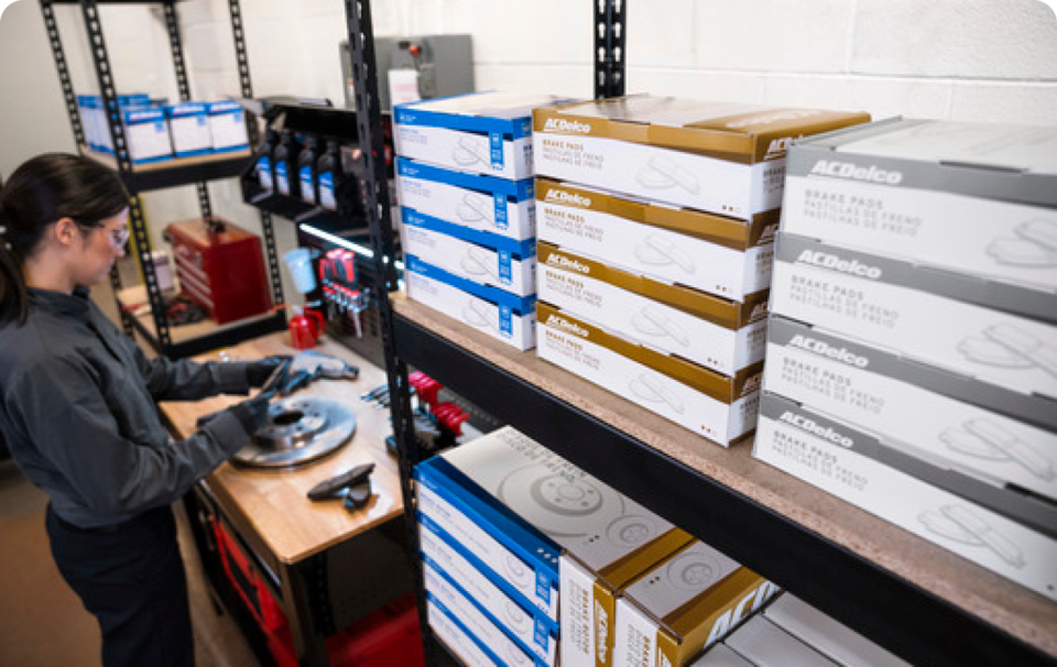 A GM Technician Inspecting a Brake Rotor Standing Next to a Shelf Full of Auto Parts